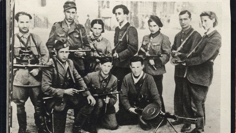 A historic black and white photo of Jewish partisan fighters standing and seated while holding firearms during wartime.