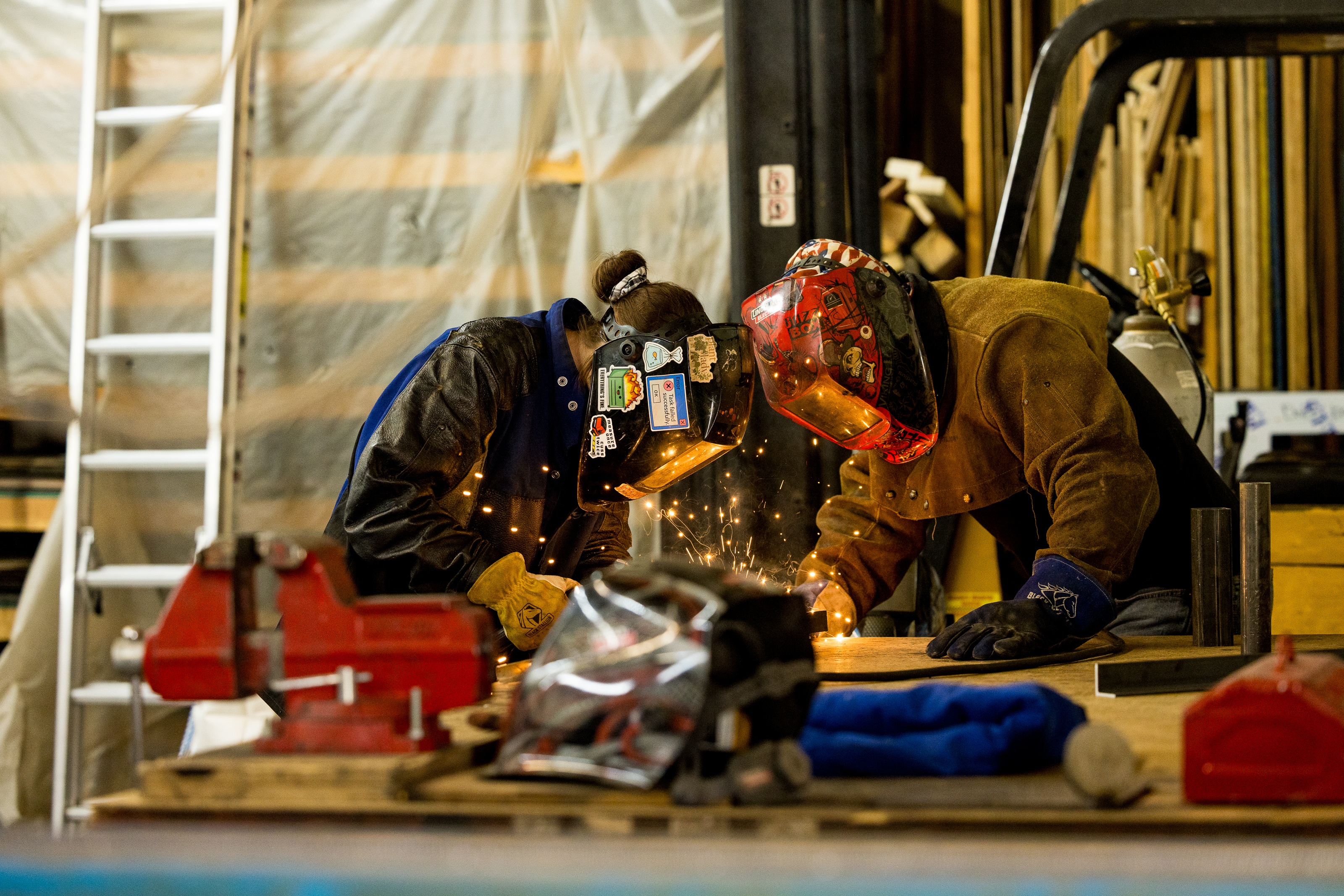 Two people wearing protective welding helmets and gloves work together on a metal project in a workshop, with sparks flying as they weld. Tools and equipment are visible on the workbench in the foreground.