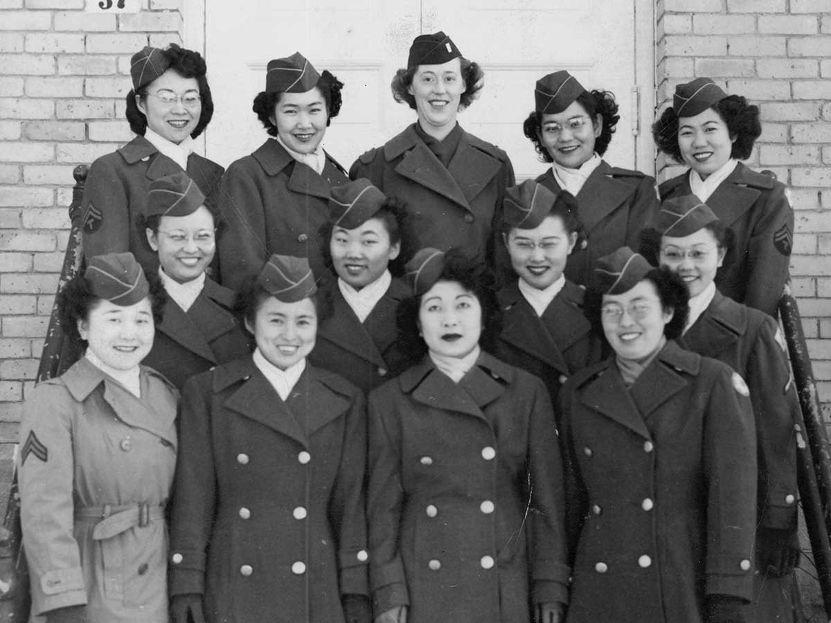 A group of women wearing matching military attire circa World War 2 stand in three rows on the steps in front of a brick building