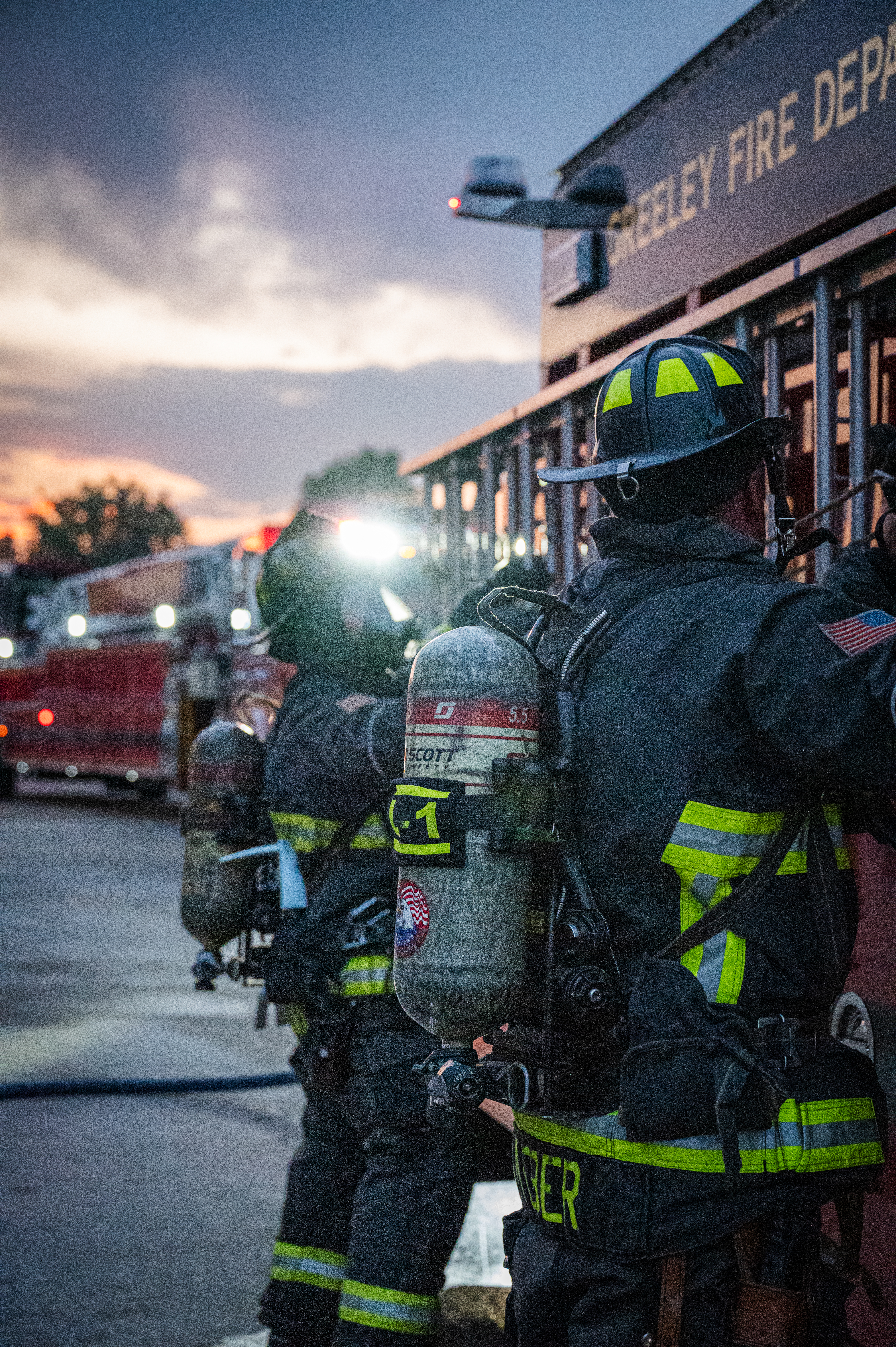 Two Greeley Firefighters in full bunker gear and helmets standing on the side of a fire truck with their helmet lights on and reaching for a ladder with a red ladder truck behind them during the evening with a sunset and cloudy background. 