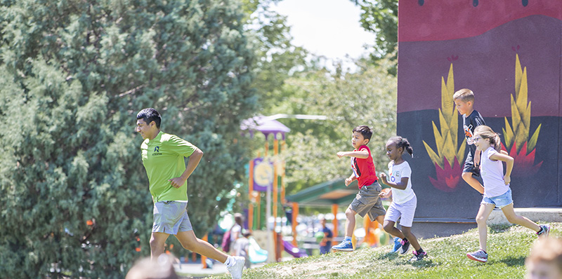 A recreation staff member leads a group of young children running down a grassy hill at Hoshiko Park on a sunny day. The kids are smiling and active, with a colorful playground and mural out of focus in the background.