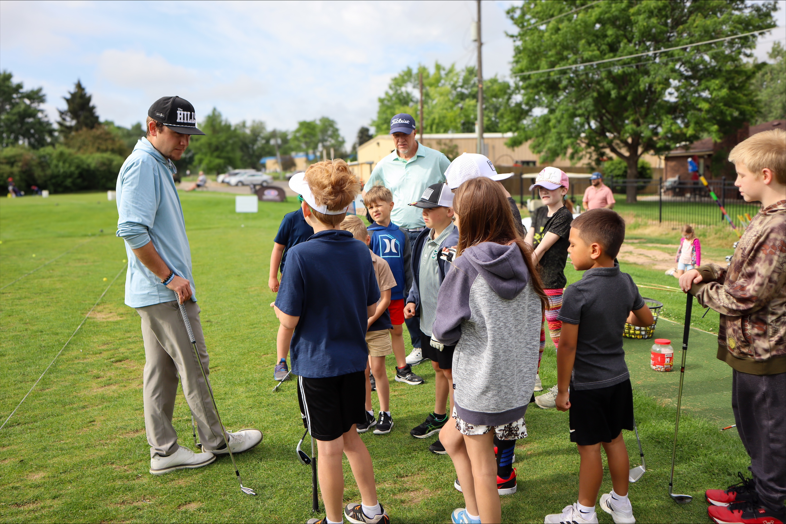 Junior golf instructors Connor Fagan and Mike Conner stand in front of a group of 12 junior golfers on the driving range, who are part of a junior golf clinic at Highland Hills Golf Course in Greeley. 