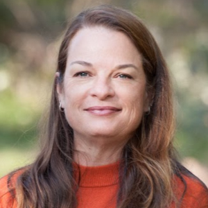 A professional headshot showing a woman with long hair wearing an orange sweater in an outdoor background.