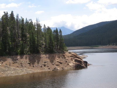 A calming view of Peterson Lake Reservoir surrounded by evergreen trees and mountains under a partly cloudy sky.