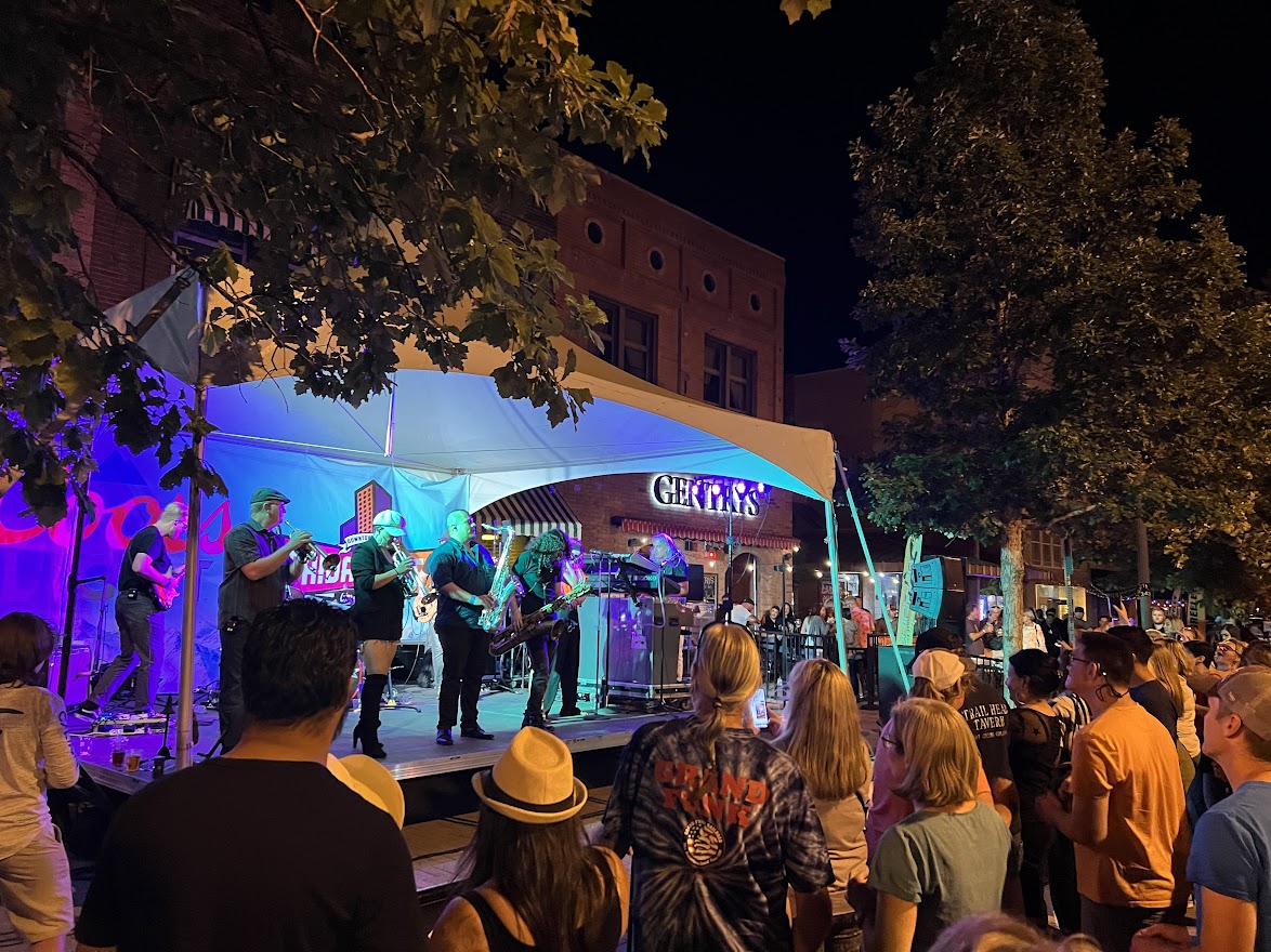 A lively evening event featuring a large crowd enjoying a live band performance under colorful lights in downtown Greeley, Colorado.