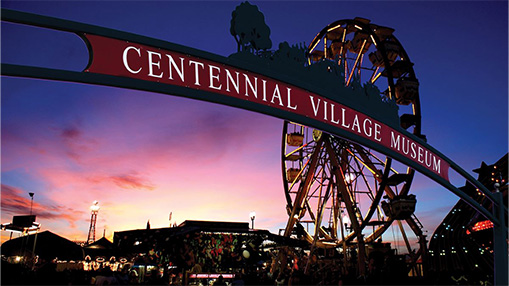 Evening view of a fairground showcasing the Centennial Village Museum with a ferris wheel and entertainment activities under a vibrant sunset.