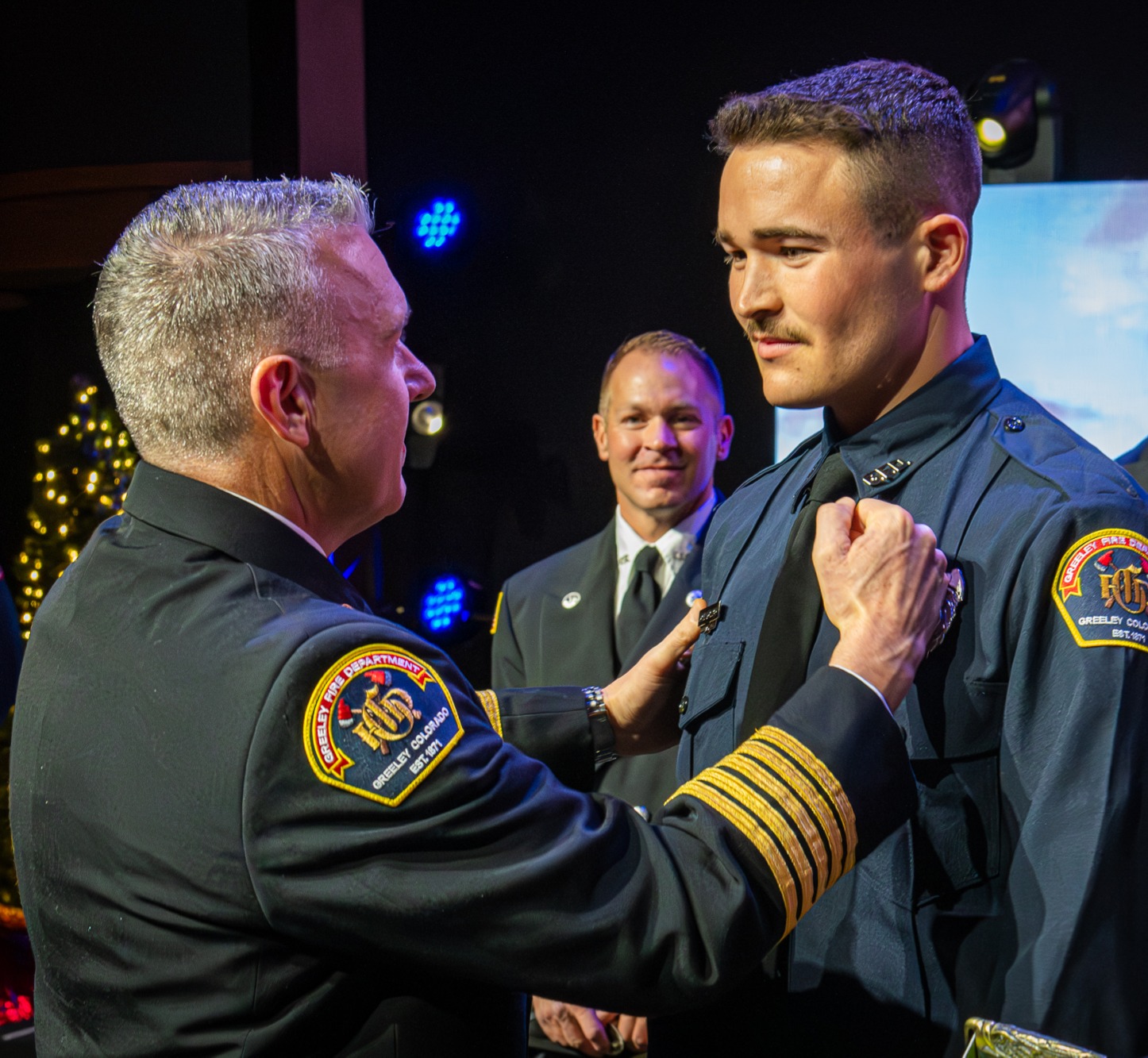 Fire Chief Brian Kuznik, in his Class A uniform, pinning a badge on a new firefighter recruit, John Bolduc, during a graduation ceremony, with someone looking on from behind. 