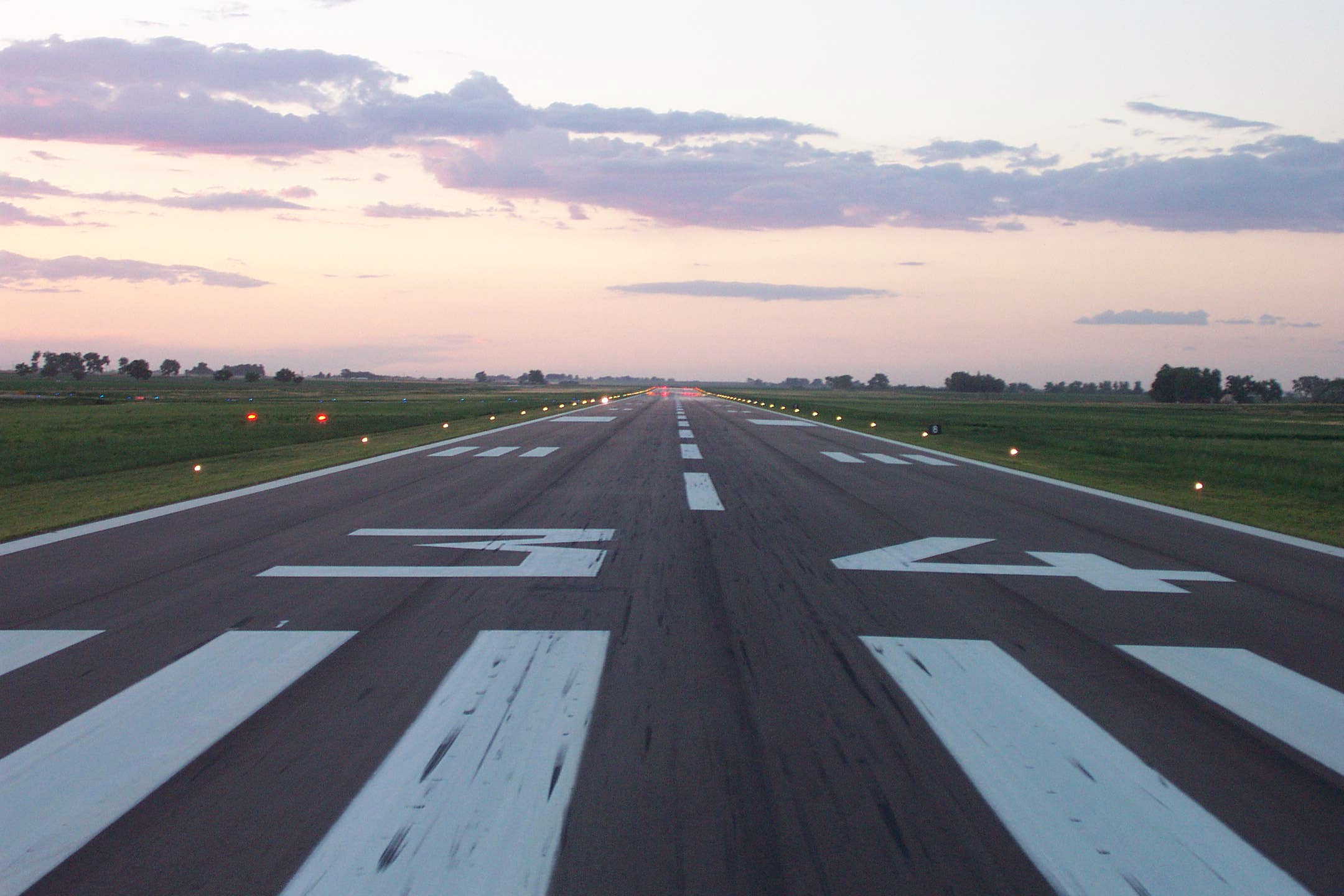 This image shows the view of runway 34 at the Greeley-Weld Airport, from the perspective of a plane landing on the runway.  In the foreground is the start of the landing area, marked by wide white paint stripes and the numbers "34".  A dotted line leads down the center of the runway.  Grass and street can be seen to the left and right of the runway, which is lined with runway lights that are lit because the sun is setting.
