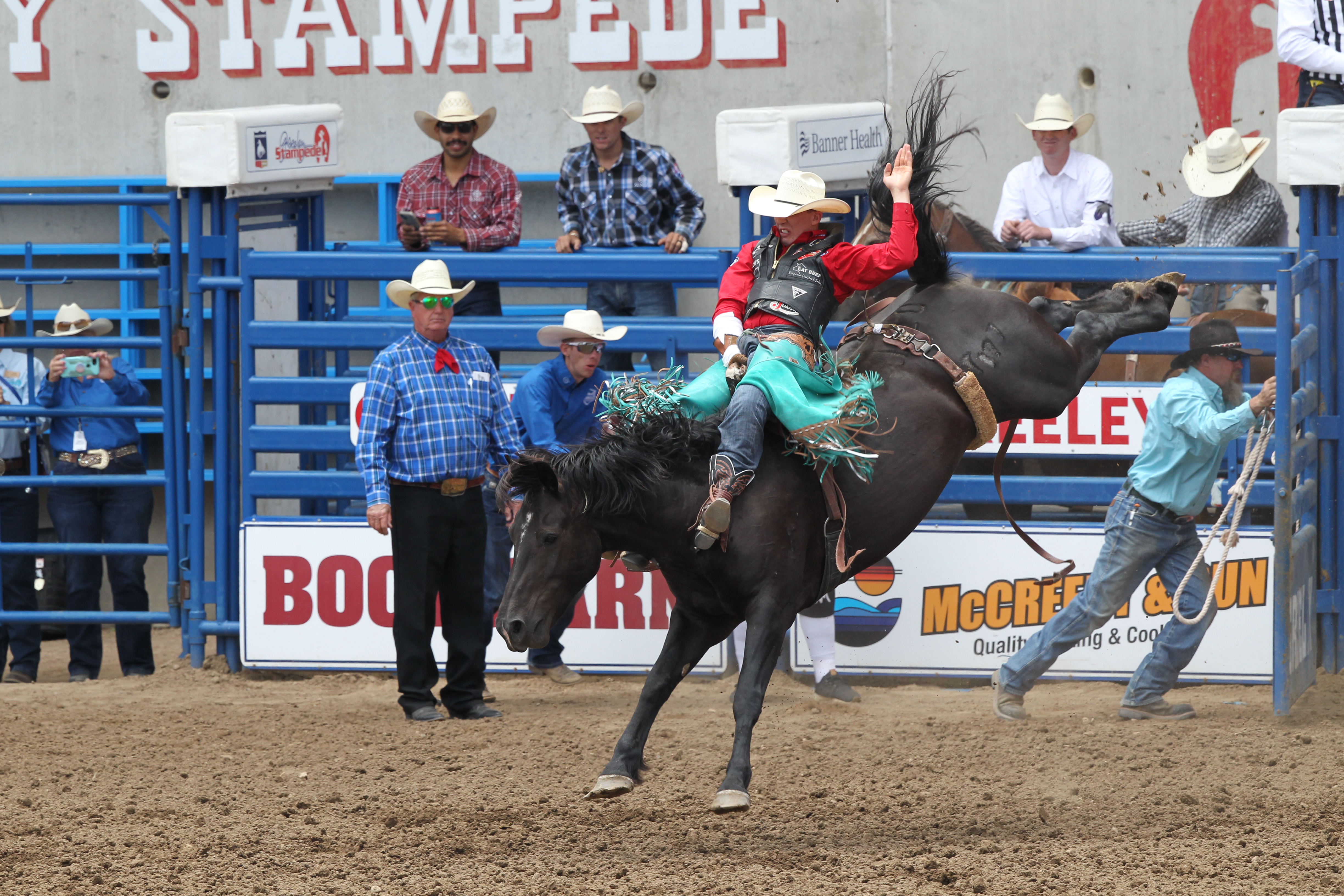 A cowboy rides and waves atop a bucking horse in an arena as spectators and officials watch in the background