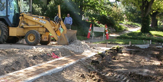 A construction crew is performing right-of-way work, using a backhoe loader to lay down gravel for street or sidewalk improvements under sunny conditions.