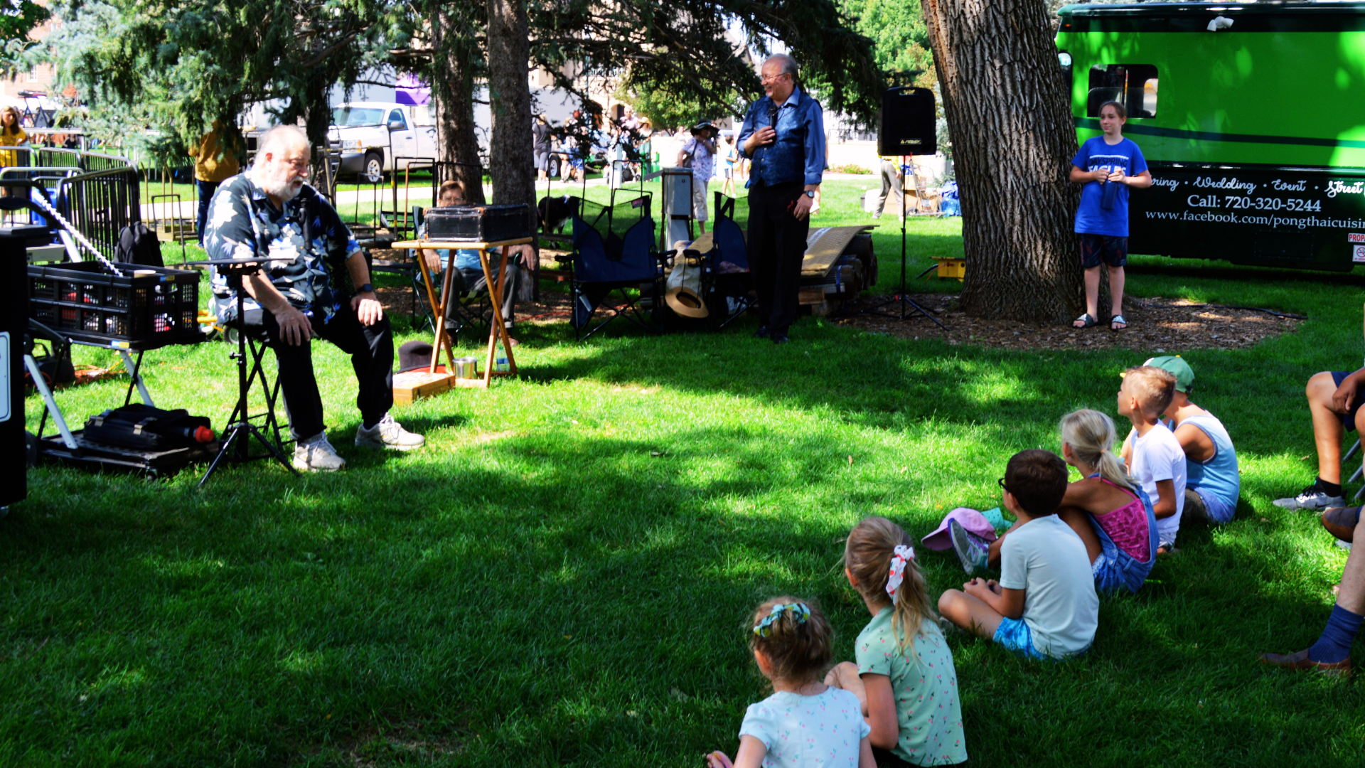 Musical act for kids' audience at the Greeley Arts Picnic. Kids sit in the grass while a musician talks about his next song. 