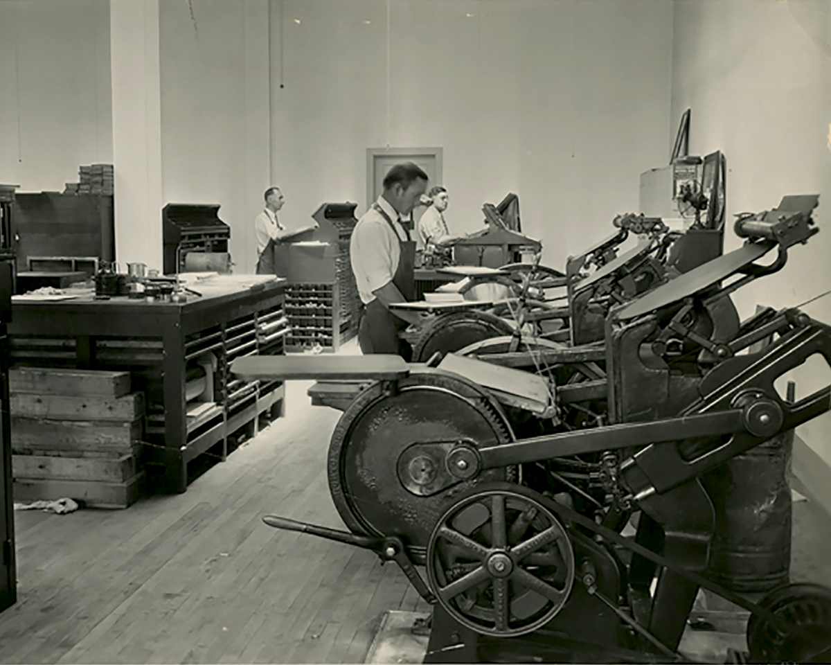Historical photo of two men standing in front of large printing presses. One man stands at a desk behind them. 