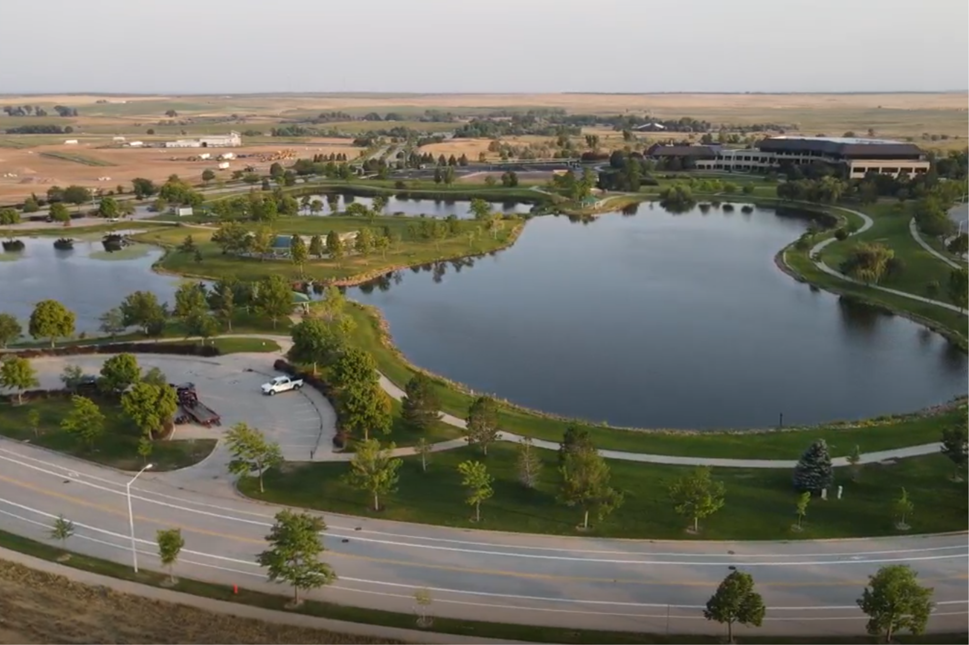 This image depicts an aerial view of JBS Foods, one of Greeley's primary employers.  The image shows a large headquarters office building in the background, with a large, serene lake taking up most of the foreground.  A road runs along the front of the property, with trees lining the road and lake.