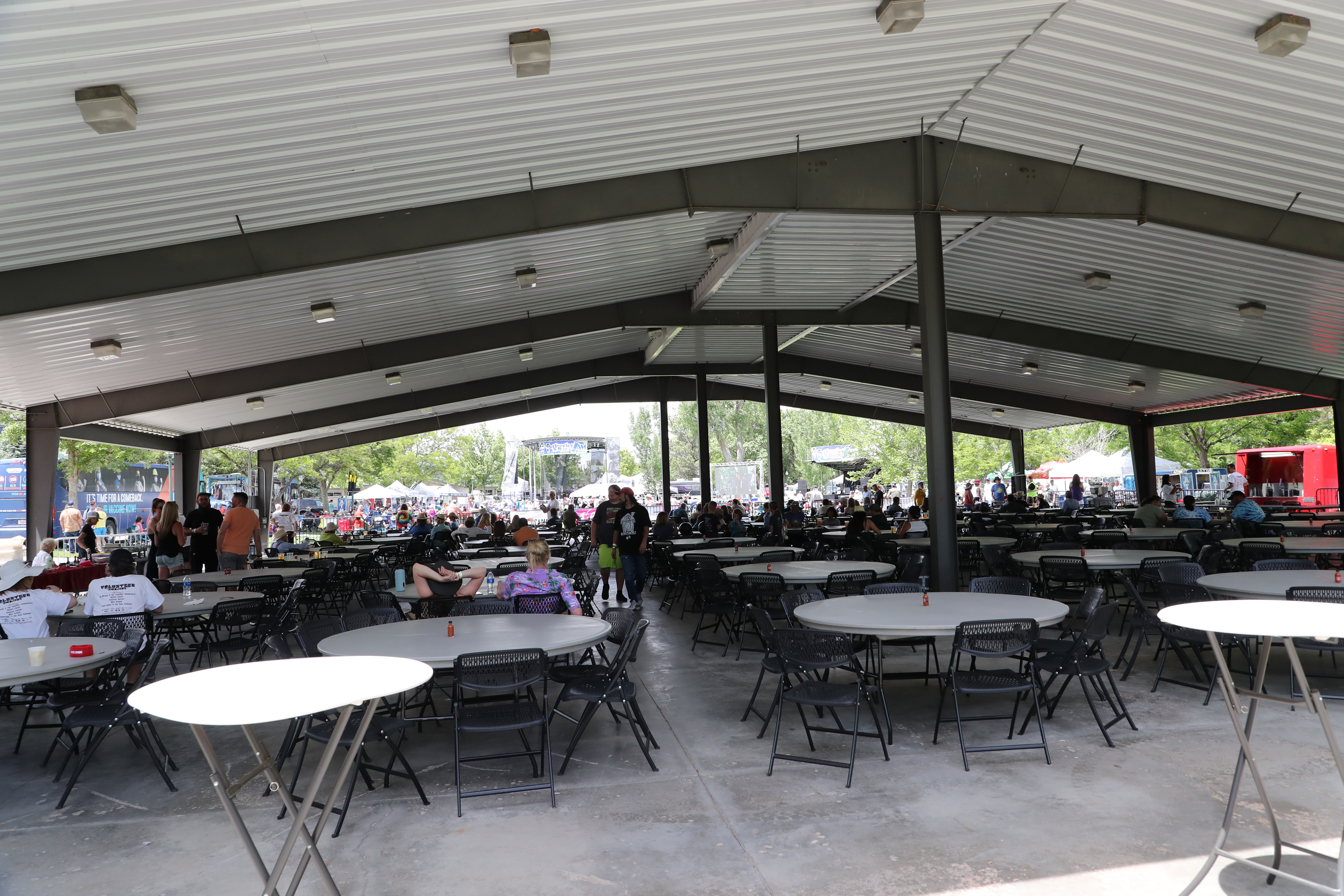 Casually-dressed eventgoers seated and standing around round tables and folding chairs beneath a large pavilion
