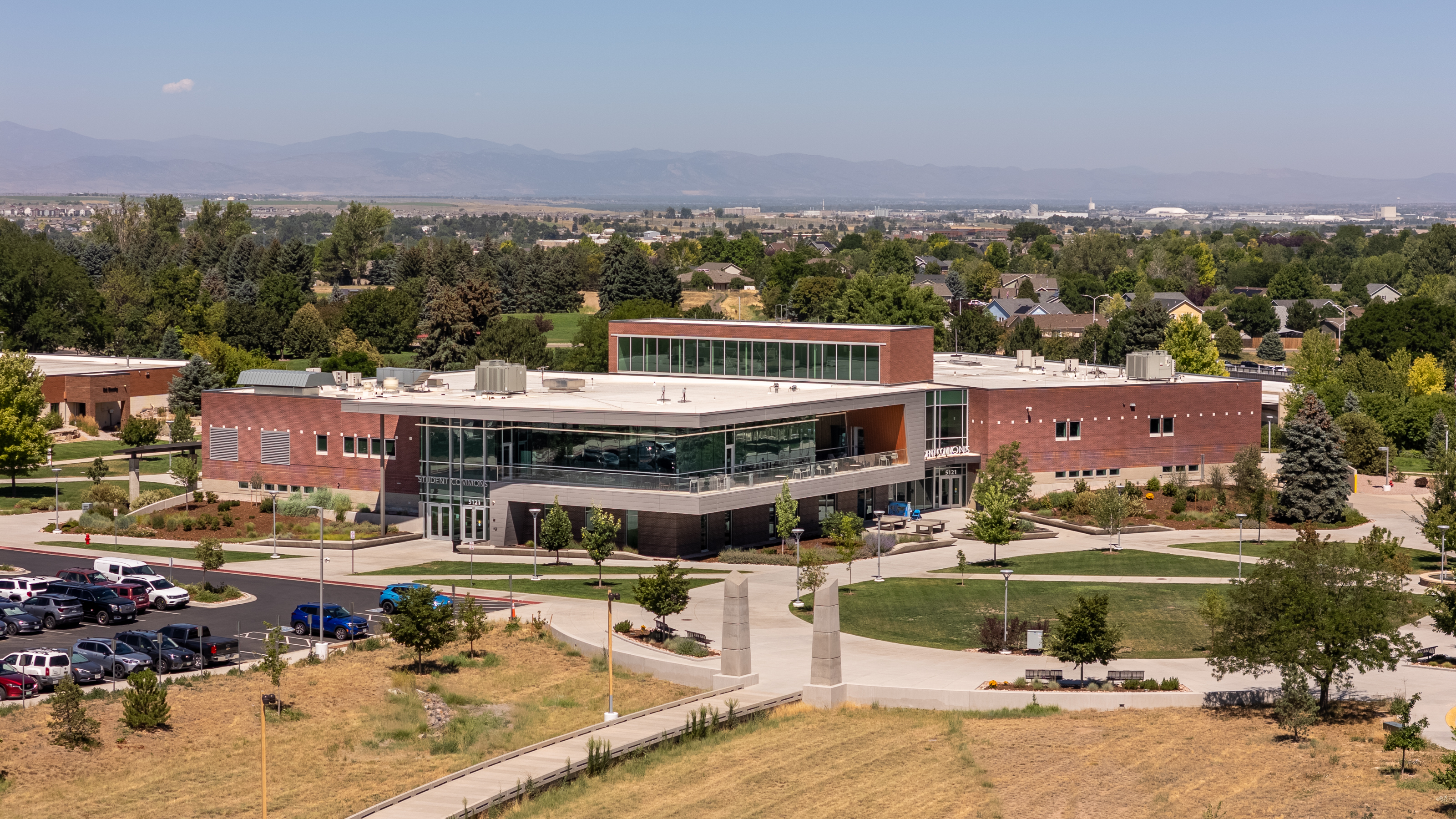 Aerial view of the Aims Community College Student Commons building in Greeley, with campus walkways, trees, parking areas, and the Front Range mountains in the distance.