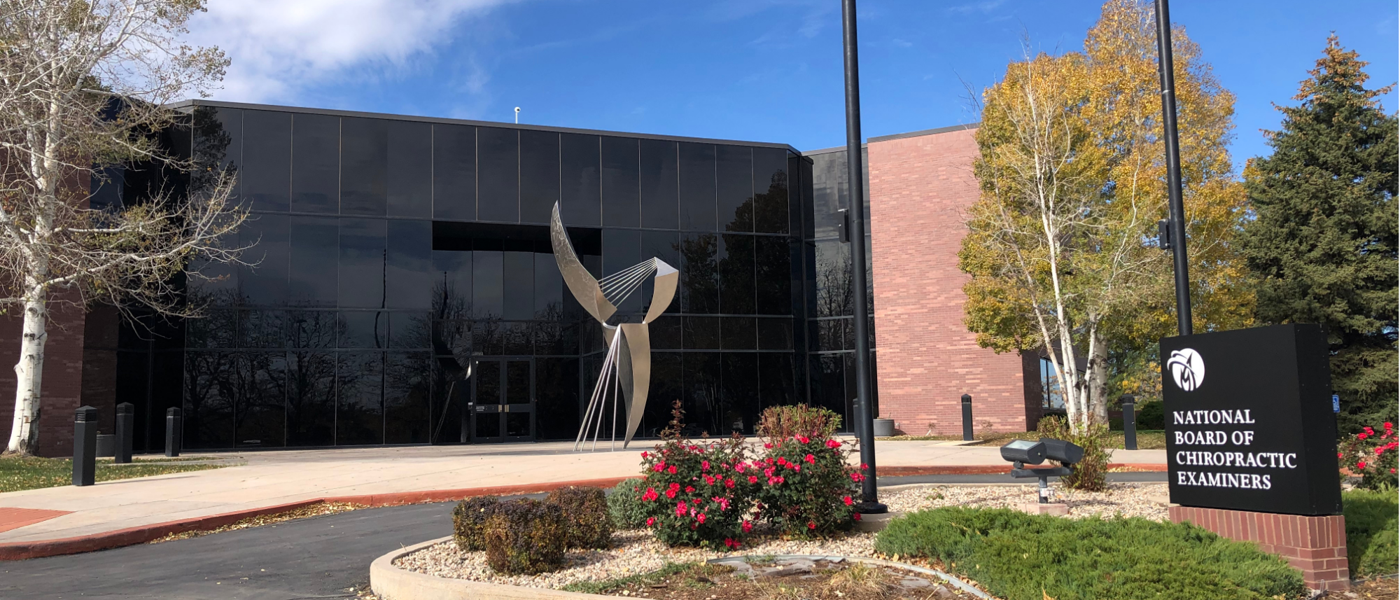 The image shows the exterior of the National Board of Chiropractic Examiners building. It features a modern architectural design with a large glass exterior and a prominent silver sculpture near the entrance. The sign in front reads "NATIONAL BOARD OF CHIROPRACTIC EXAMINERS" against a dark background. 