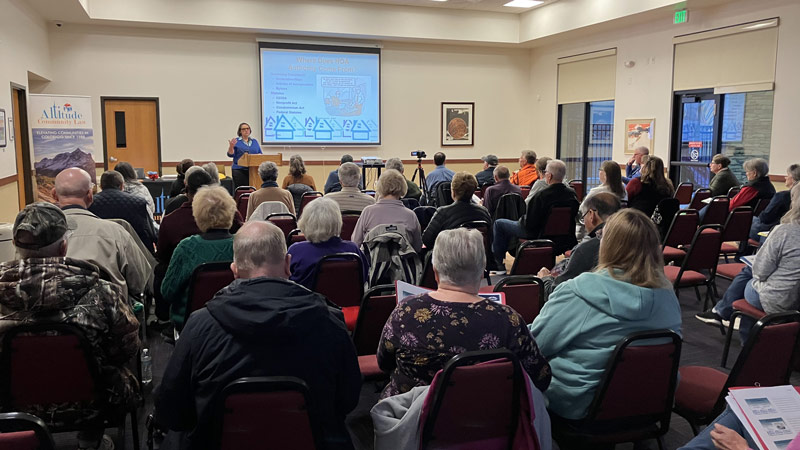 A woman at a podium speaks to a room filled with engaged attendees during a community event.