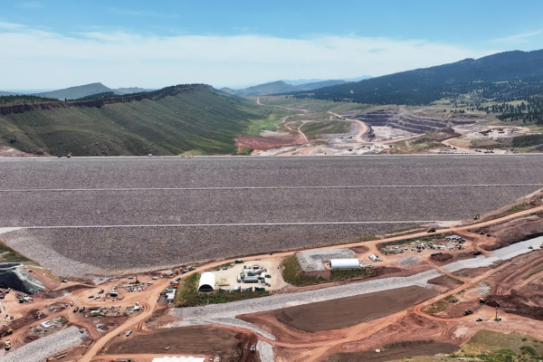 A wide aerial view of Chimney Hollow Dam in Colorado, showing the completed rock-fill dam spanning a valley, with construction roads, equipment, and terraced excavation areas in the foreground and mountains in the background.