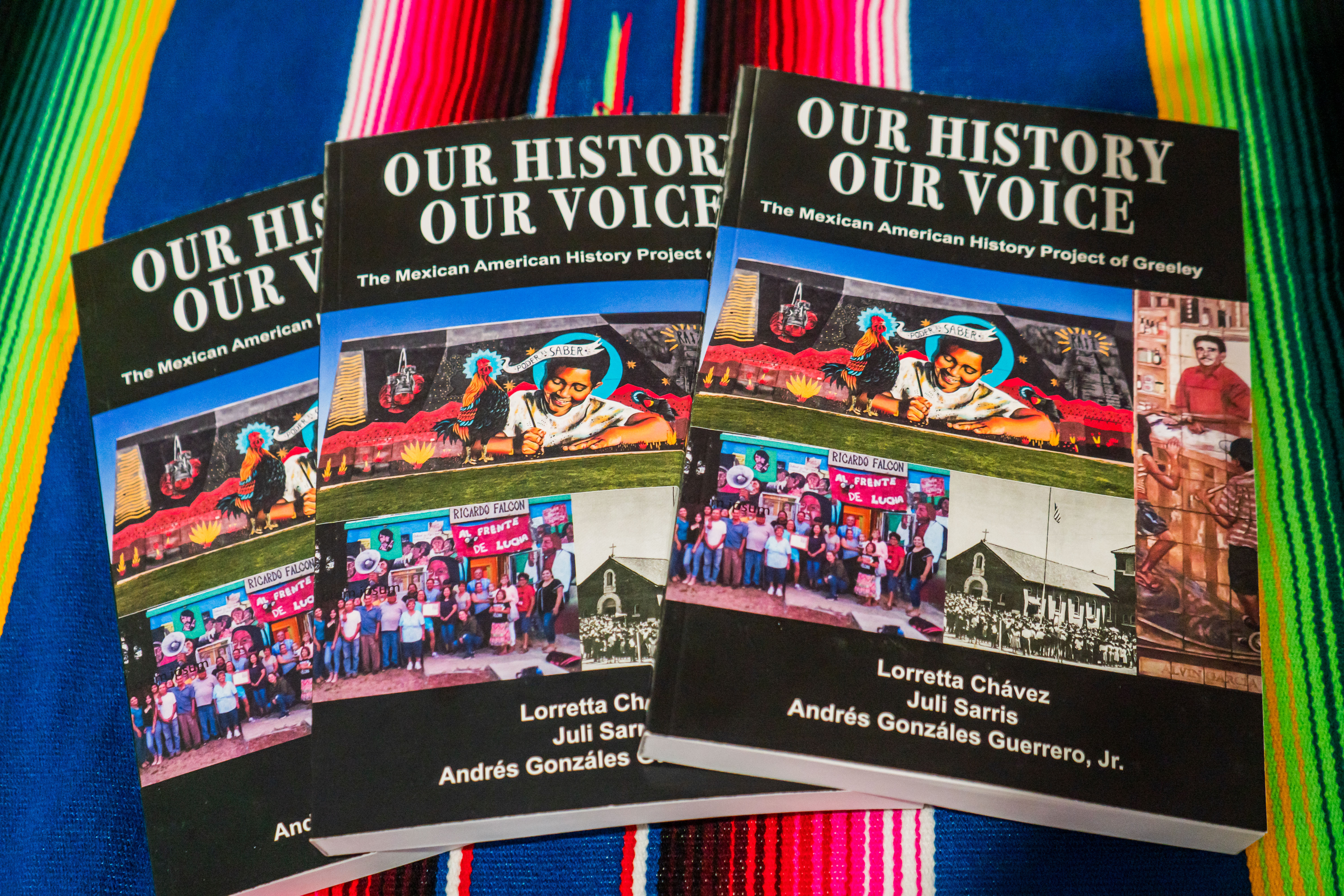 Three copies of a paperback book titled Our History, Our Voice: The Mexican American History Project of Greeley sit on a table covered by a sarape.