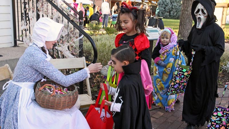 Children in costumes hold out bags to collect candy from a costumed individual on the porch of a historical building.