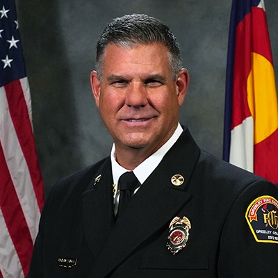 Professional photo of Greeley Fire Department's Battalion Chief Richard Waidler in his Class A uniform with the US and Colorado State Flag behind him. 