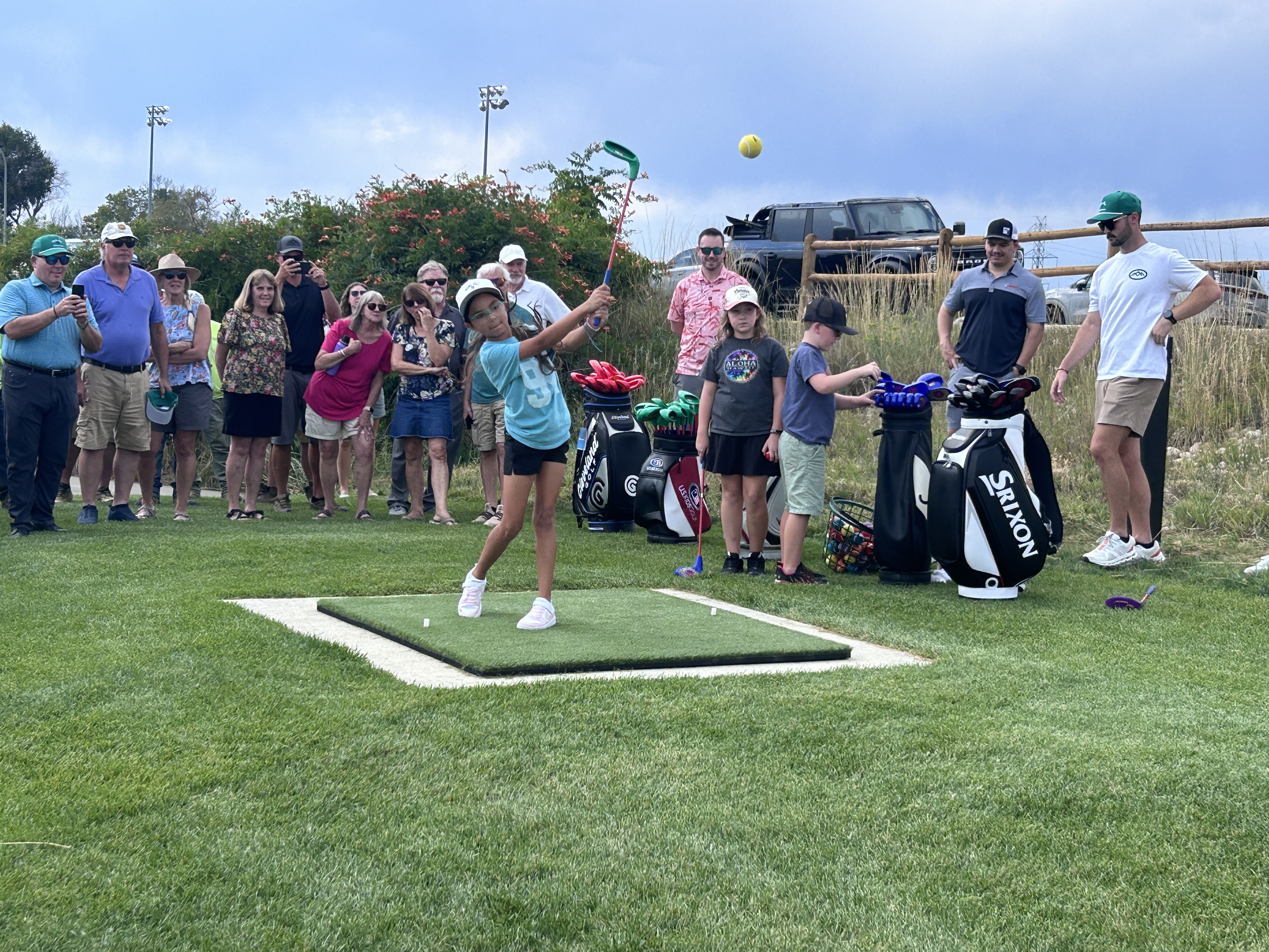 Junior golfer swinging a golf club on a turf mat at an opening ceremony while a crowd watches in the background.