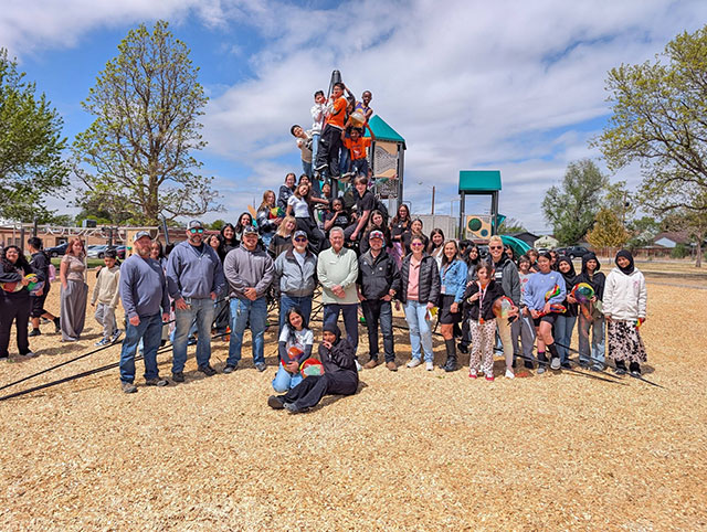 A group of people posing around a new playground, with kids climbing the structure