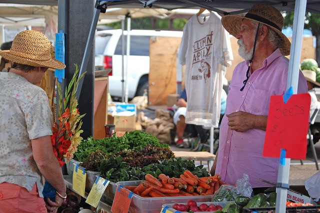 A vendor interacts with a customer at a farmer's market stand offering fresh vegetables and flowers.