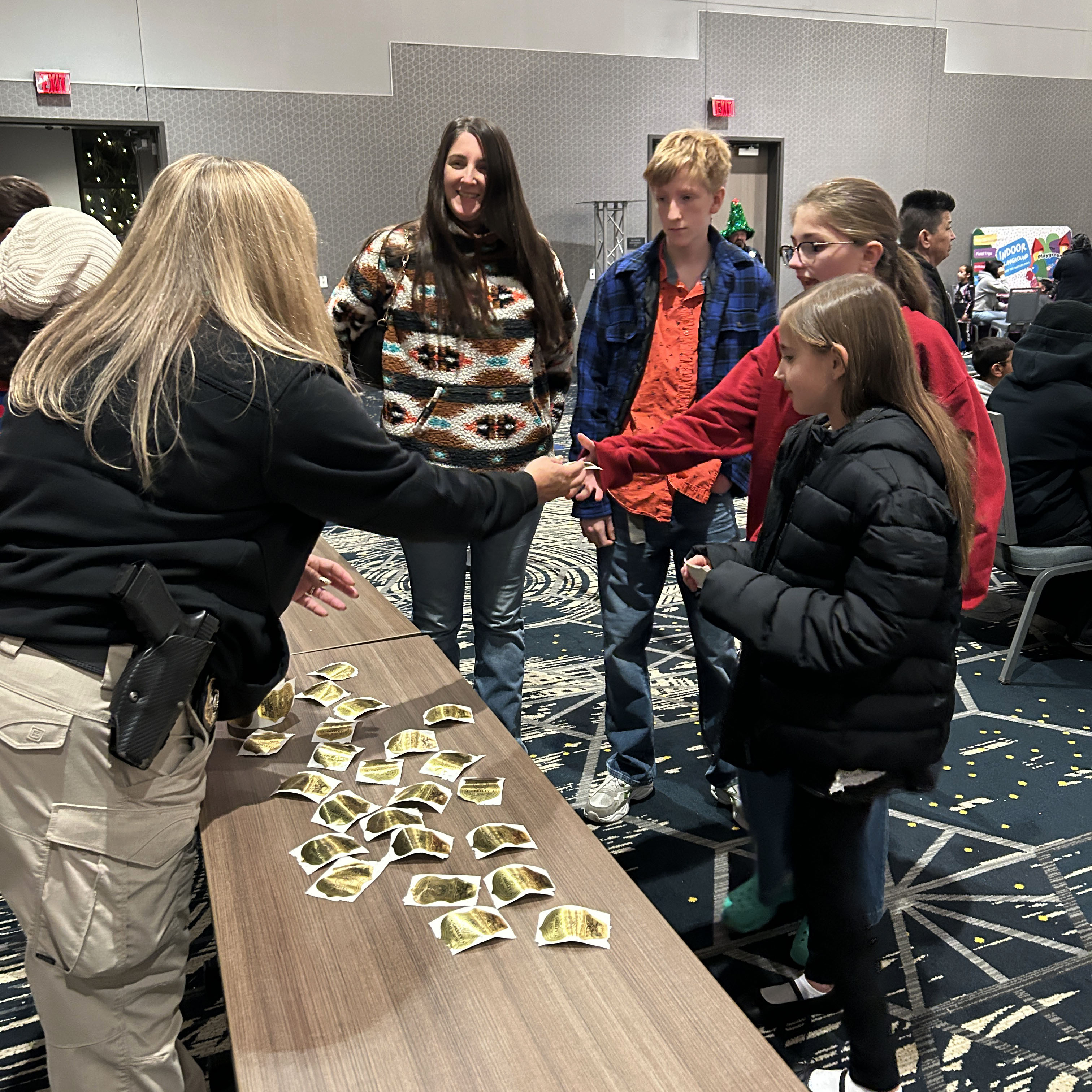 A Greeley Police Detective hands out badge stickers to families during the GPD's annual gift-giving event. The officer is wearing a black jacket and khaki pants, leaning over a table with stickers on it, handing to a girl wearing a red sweatshirt. There is a girl in a black jacket in front of her, and a boy in a blue and black plaid shirt as well as a woman in a colorful sweater behind her.