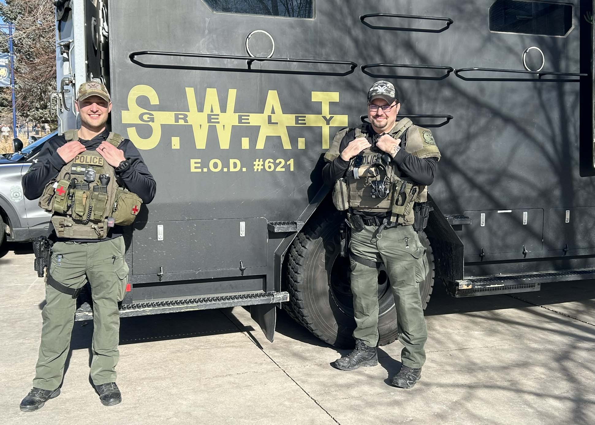 Two officers with the Greeley Police Department pose in front of a black SWAT truck. The officers are wearing bullet-proof vests and baseball caps and are smiling at the camera.