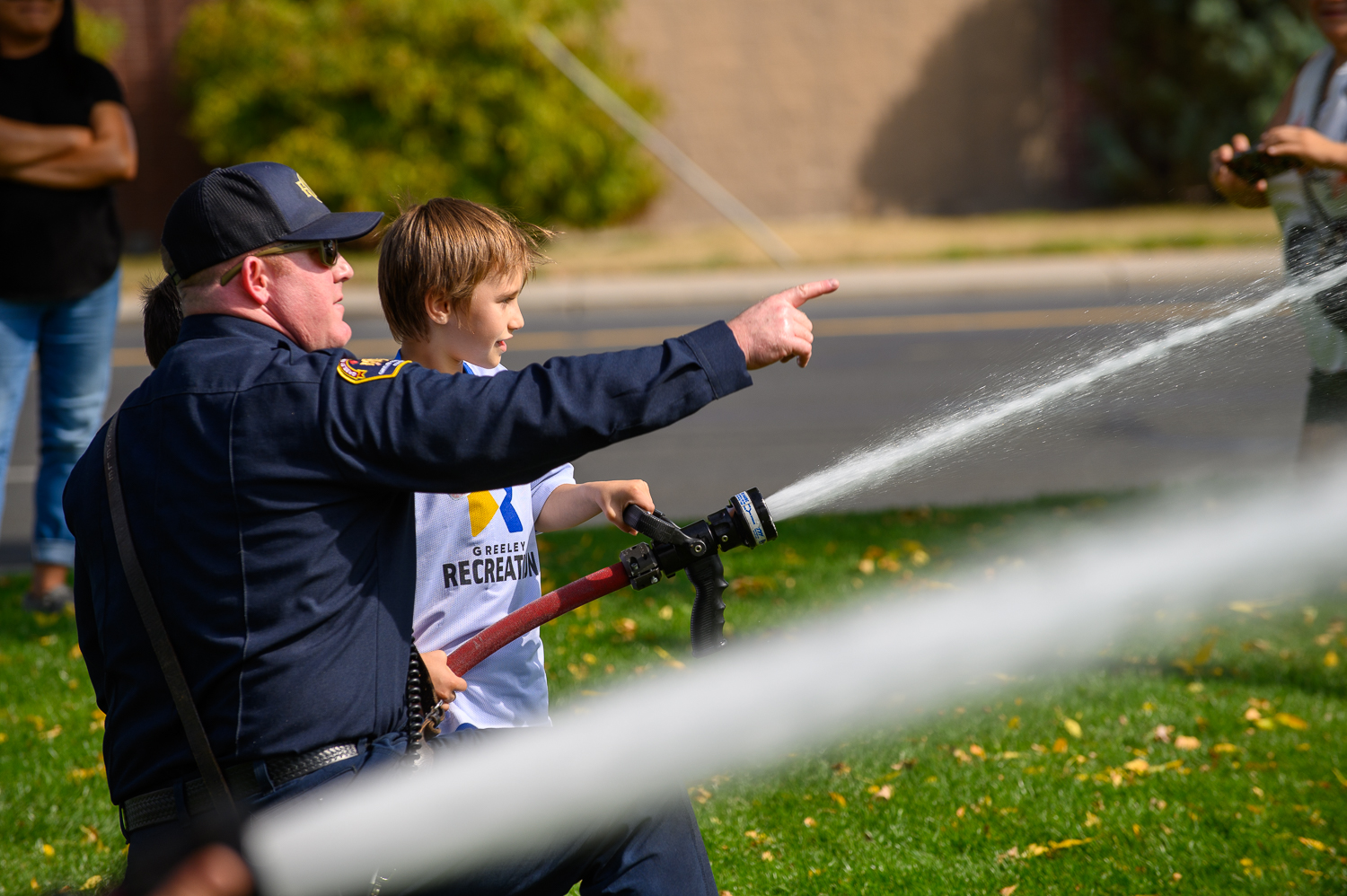 A Greeley firefighter points his finger to the right, directing a child beside him spraying water from a fire hose. 