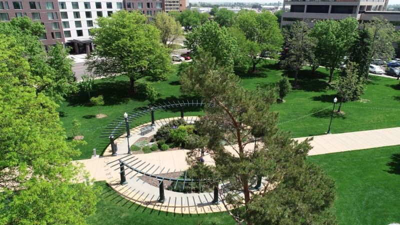 An aerial view showcasing a curved walkway adorned with decorative metal archways, situated in a vibrant urban park. The scene highlights lush green lawns, mature trees, and nearby city structures.