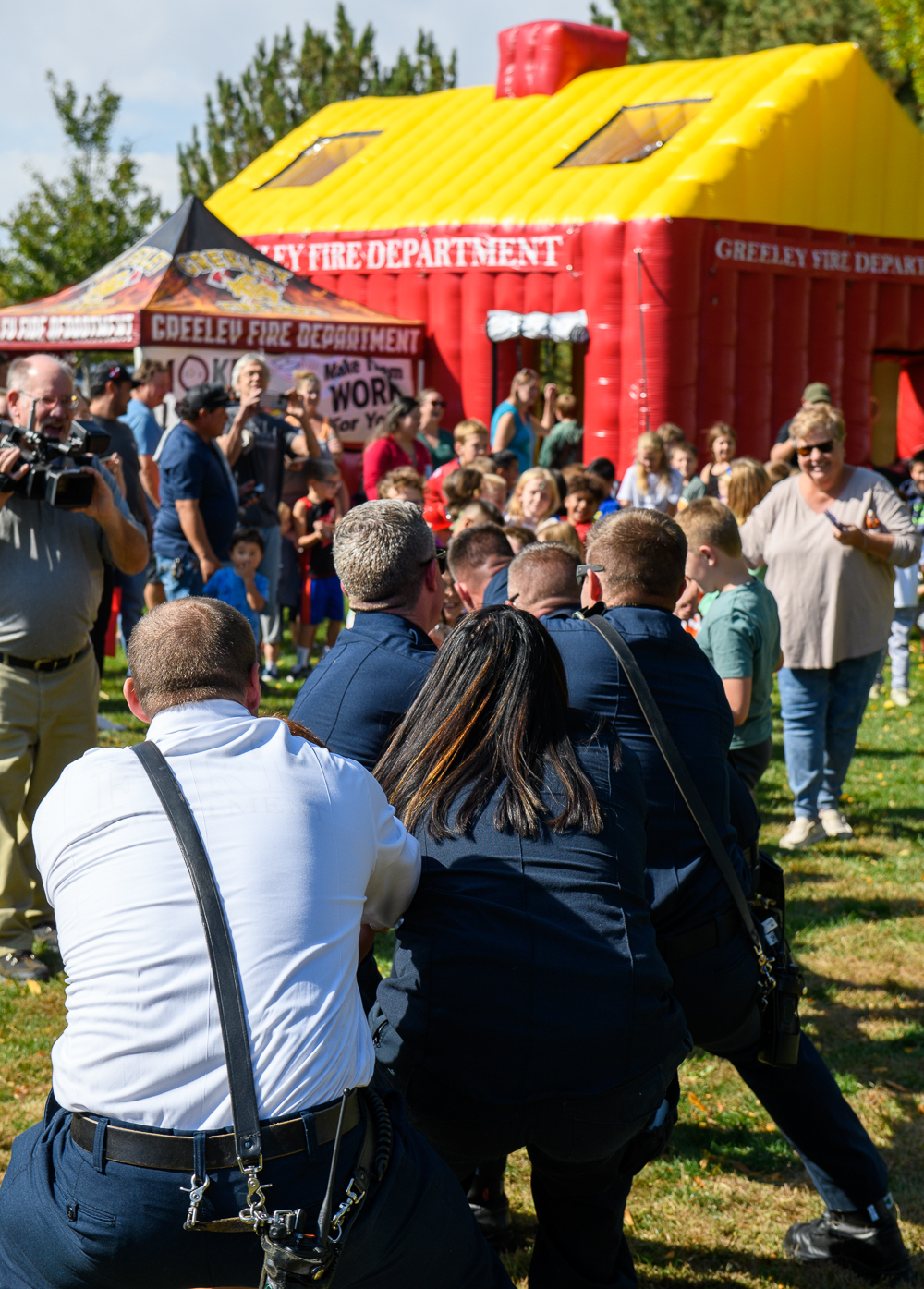 The backs of several Greeley firefighters are seen as they play tug of war with children at Greeley Fire's annual Open House. An inflatable house, canopy, and crowd can be seen behind the participants in the game. 