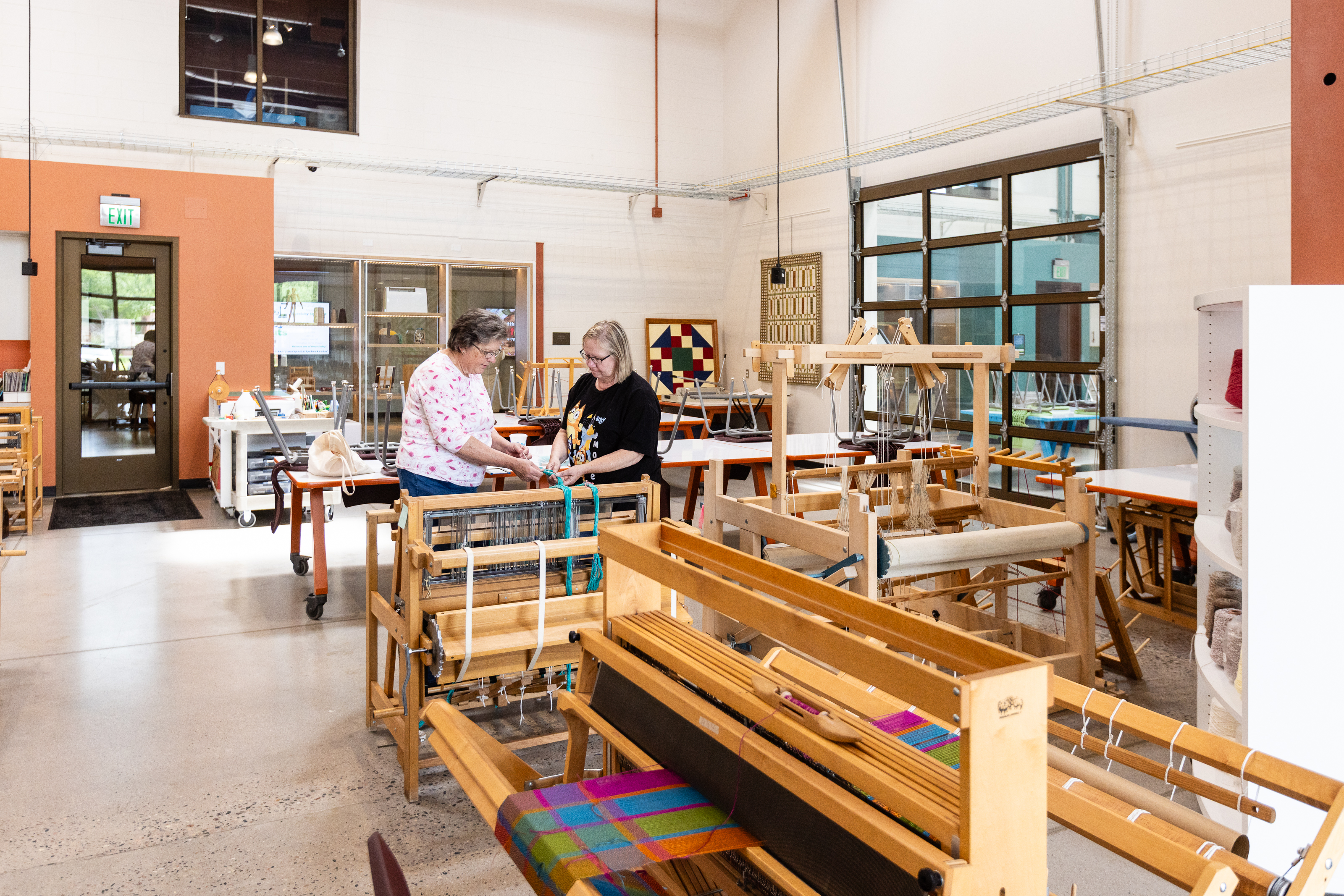 Two people work at a weaving loom inside the LINC Library Innovation Center textile studio, surrounded by looms, craft materials, and classroom equipment.
