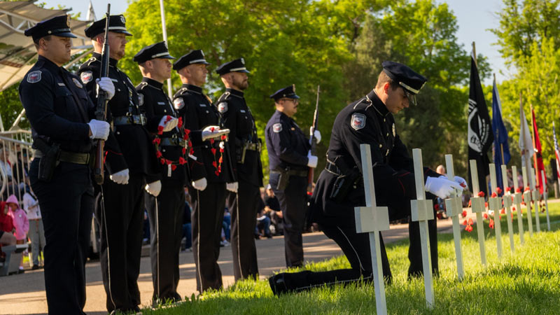An officer kneels to place a red poppy wreath on a white cross grave marker during a ceremonial event.