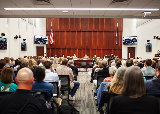 A City Council meeting with the council seated at the front of the room and a crowd of community members seated throughout the room