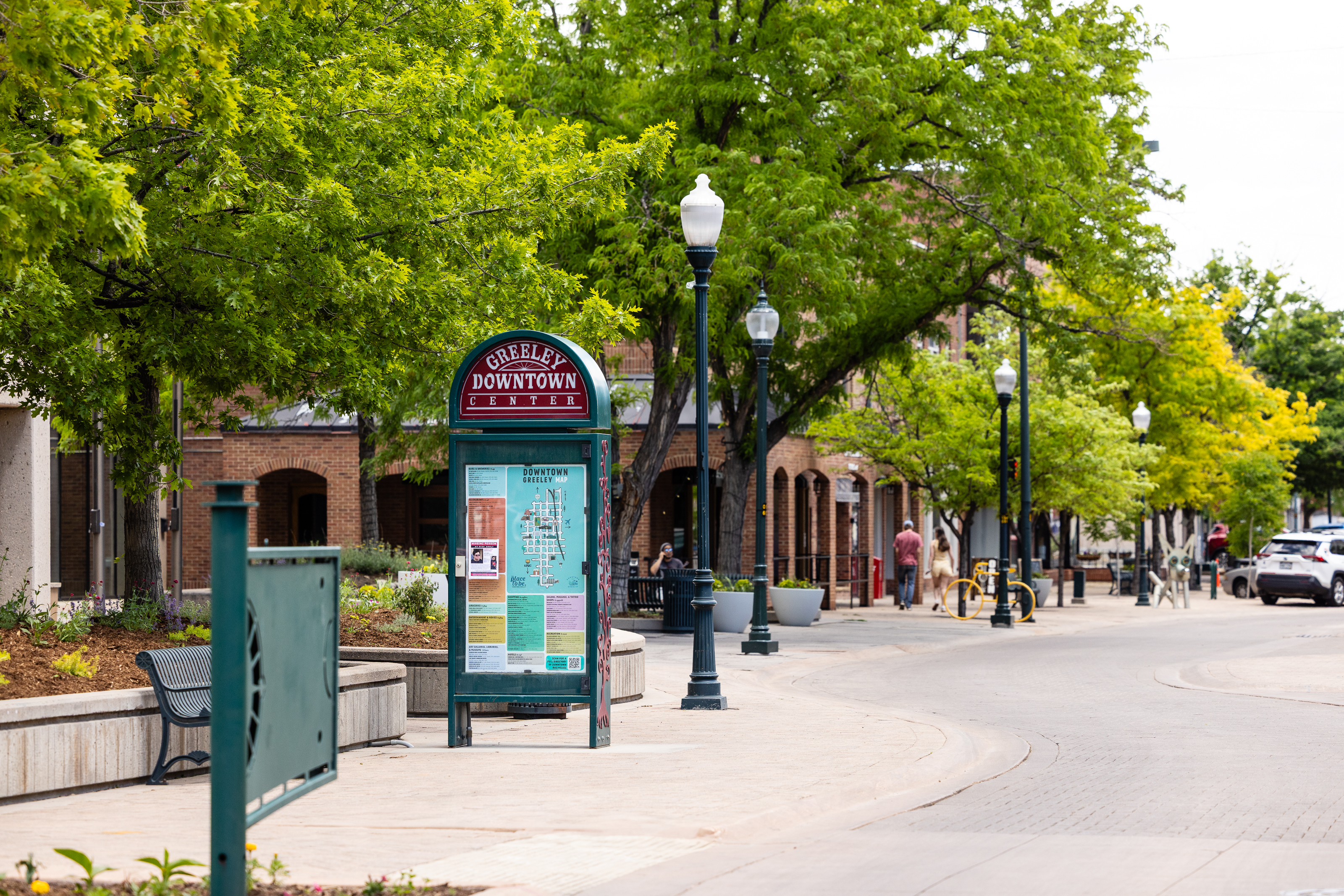 This image depicts downtown Greeley, Colorado.  A large brick building with archways can be seen behind lush green trees and a sign that reads "DOWNTOWN GREELEY".  Quaint streetlights line the sidewalk, which curves along 8th Avenue in downtown Greeley.  
