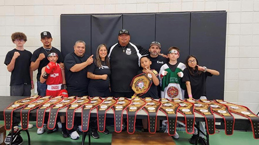 A group of individuals celebrates boxing achievements in a community center, showcasing numerous awarded belts.