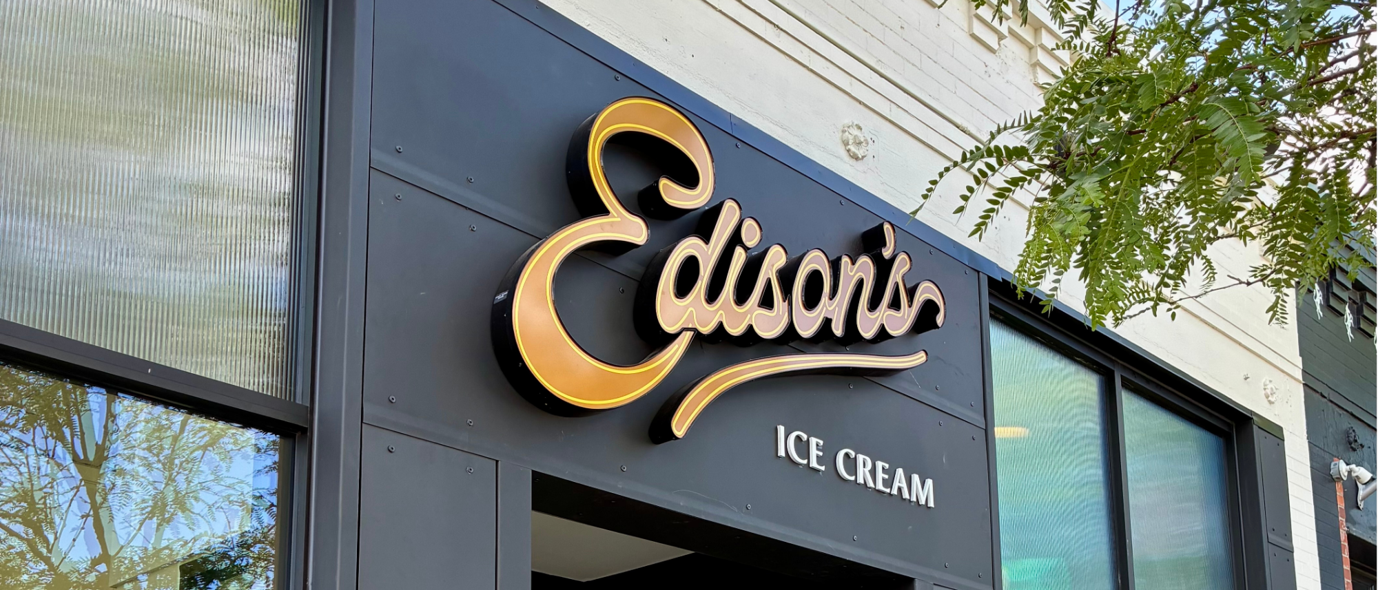 Exterior of Edison’s Ice Cream in downtown Greeley, featuring a black storefront with a gold script “Edison’s” sign above white “Ice Cream” lettering, framed by windows and tree branches.