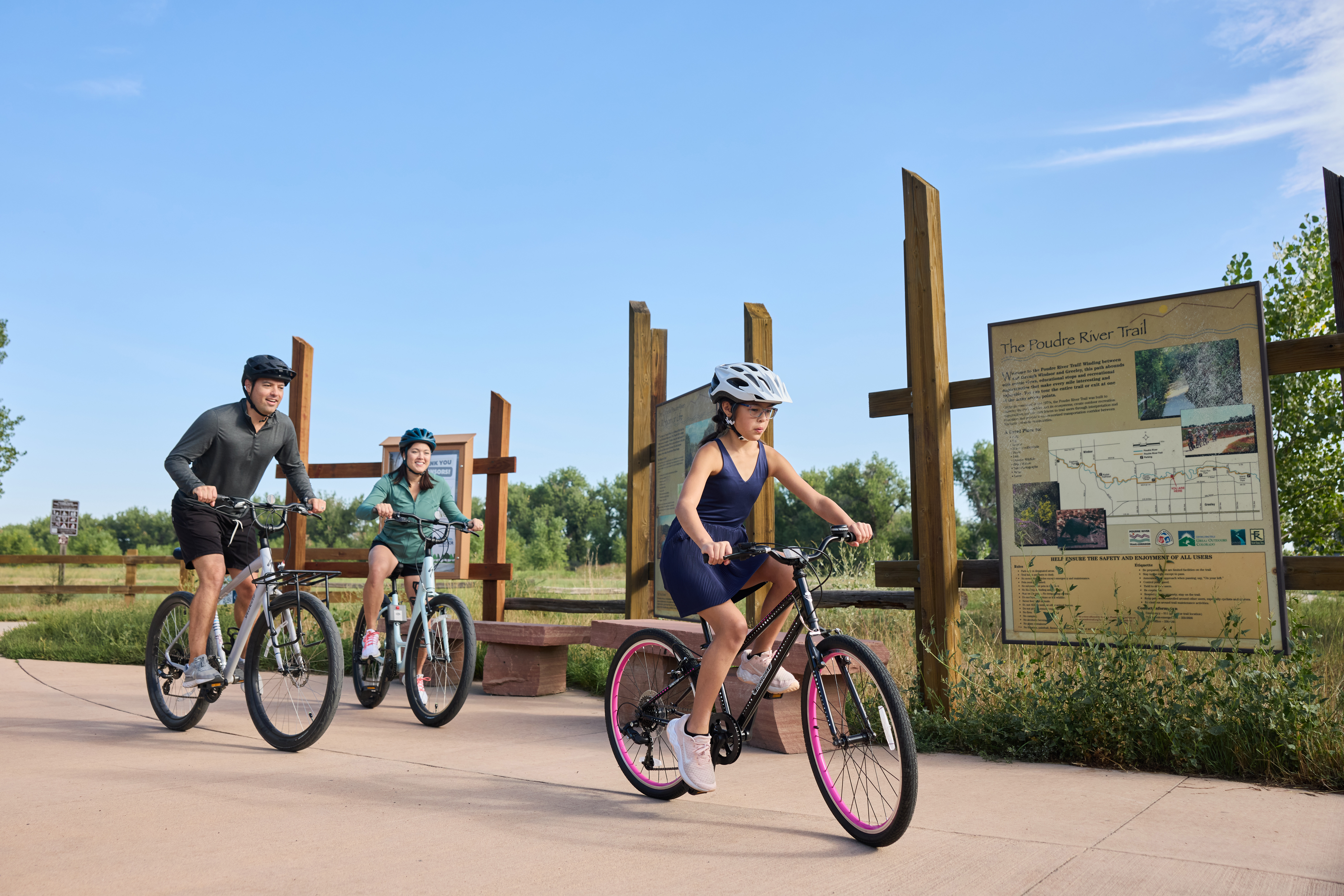Two adults and one child are riding individual bicycles on a concrete trail.  The child is in the lead and is riding a black bicycle with pink tire rims.  The child is wearing a blue sleeveless jumper and white helmet.  The adults are riding side-by-side behind the child.  The man is riding a white bicycle and is wearing black shorts, a green shirt, and a black bicycle helmet.  The woman is riding an aqua blue bicycle and is wearing black shorts, a green jacket, and a blue bicycle helmet.  The family is biking past three large signs on wooden posts which provide maps and information about wildlife on the Poudre River Trail.