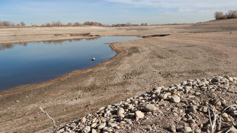 A dry, barren pond with a small patch of water remaining near the center, surrounded by sandy and rocky terrain.