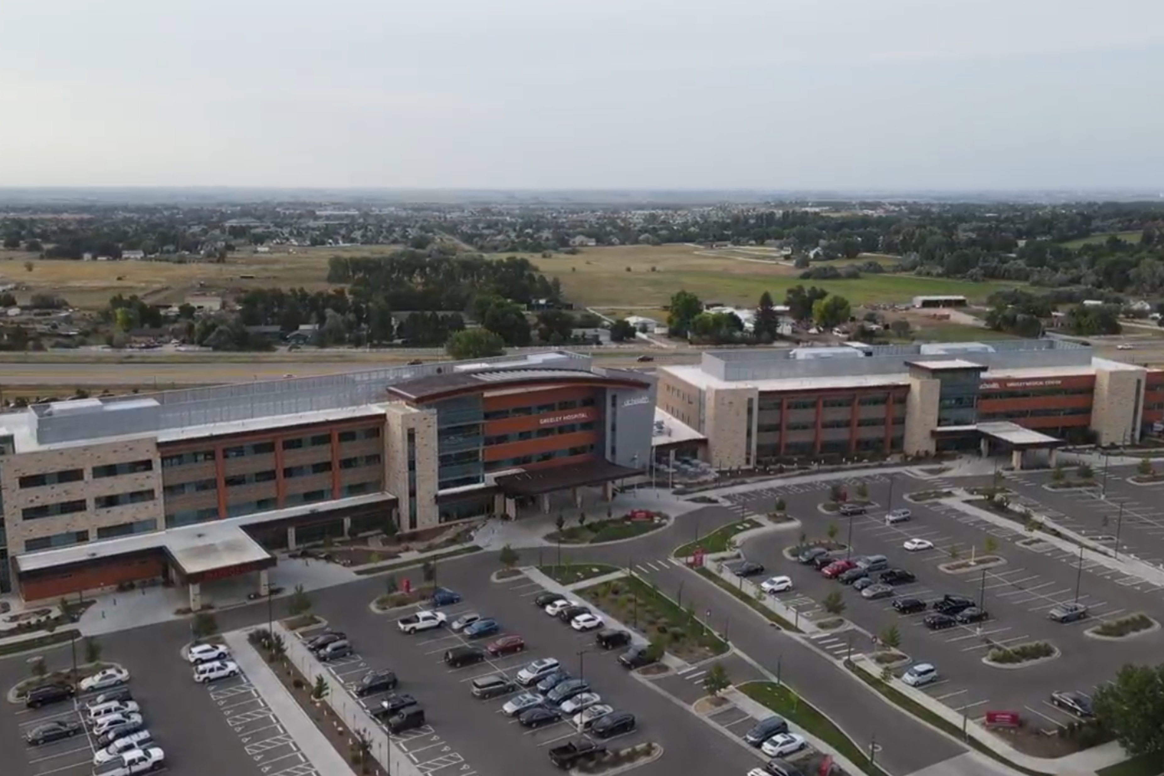 This image depicts UC Hospital in Greeley, Colorado.  An expansive, modern, four-story hospital building is centered in the photograph.  The building facade is a combination of glass, tan stone, and burnt orange stucco.  A large parking lot spans the length of the building and is peppered with parked vehicles.  Acres of large grassy areas and trees can be seen behind the hospital building.