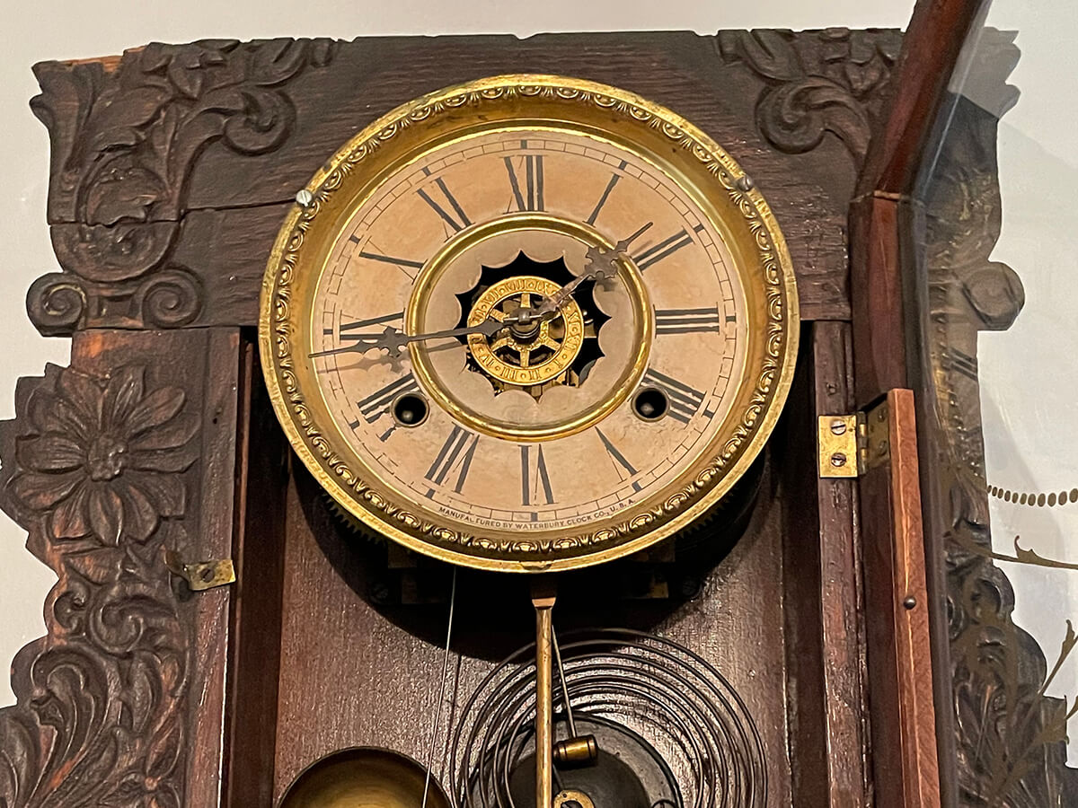 Closeup of an ornate, antique clock with roman numerals and a pendulum, mounted on an intricately carved wood frame