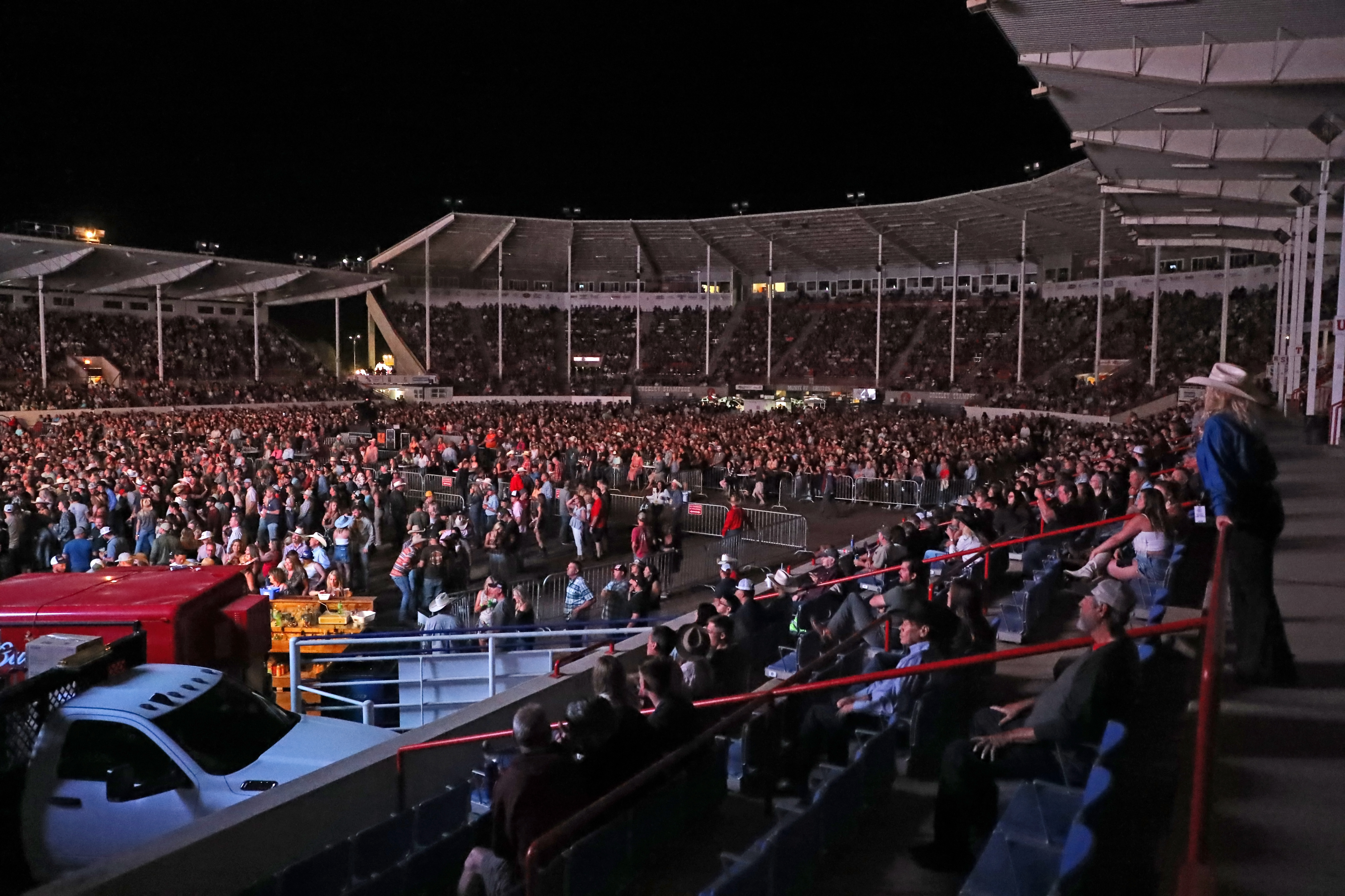 A large crowd of seated and standing spectators at the Concert View Arena gather at night during the Greeley Stampede