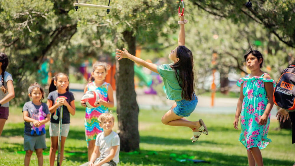 One young girl attempts the slack line challenge as she swings through the air while other children look on during a Play on the Way outdoor activity at the park