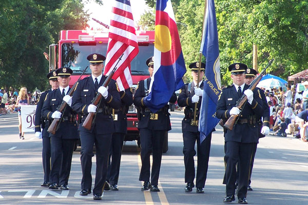 A group of police officers marching in a formal parade holding flags, with a fire truck and crowd in the background.