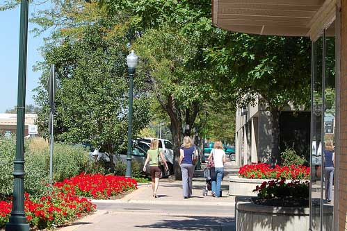 A vibrant sidewalk lined with trees, flowers, and retail surroundings depicting community engagement.