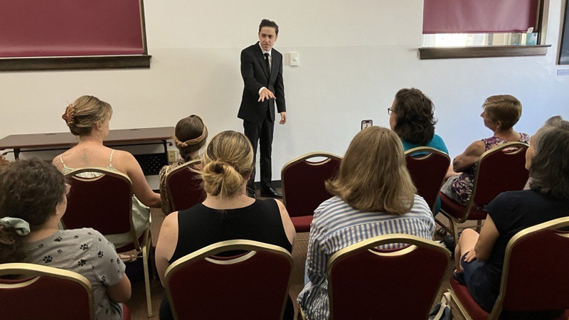 A young presenter in a black suit delivers a talk to an engaged audience in the community room of the Greeley History Museum.