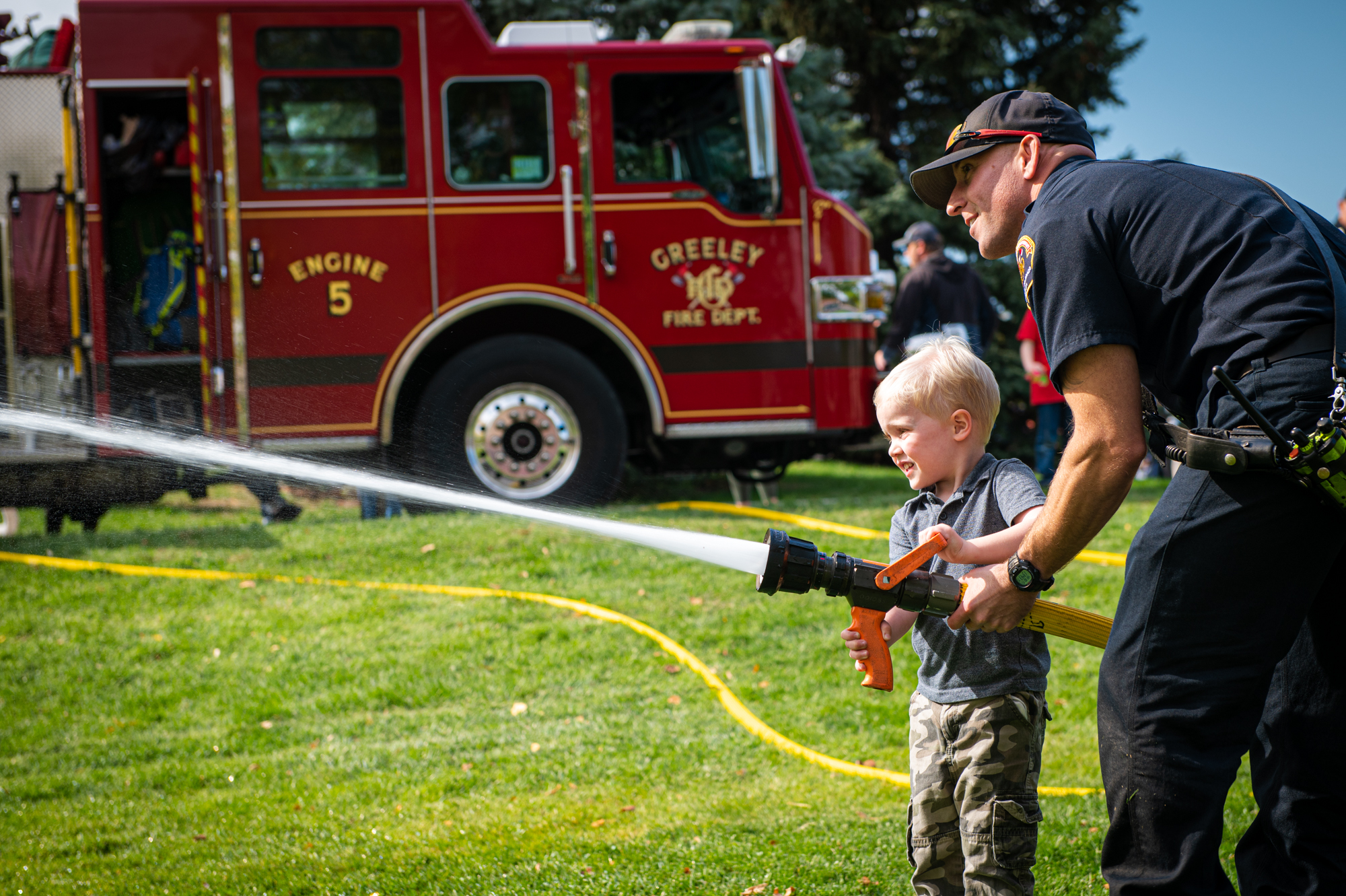 A child is seen holding a fire hose that is spraying water with the assistance of a Greeley Firefighter. 