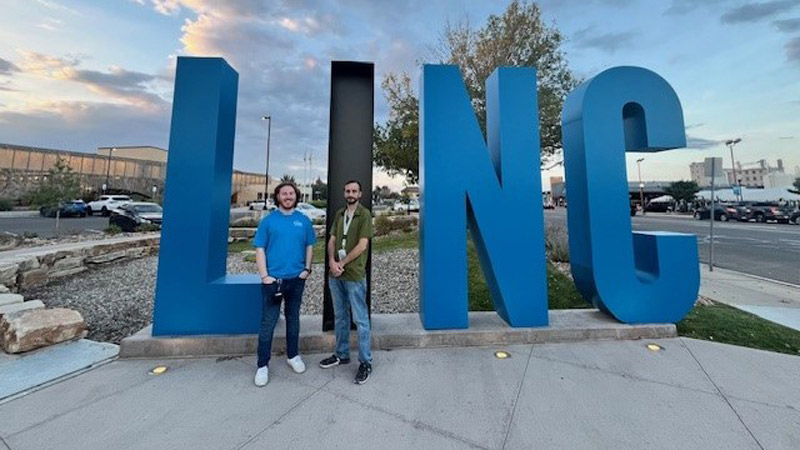 Two men stand near a large oversized sign that reads LINC in blue and black letters, with cars, buildings, and a parking lot visible in the background.